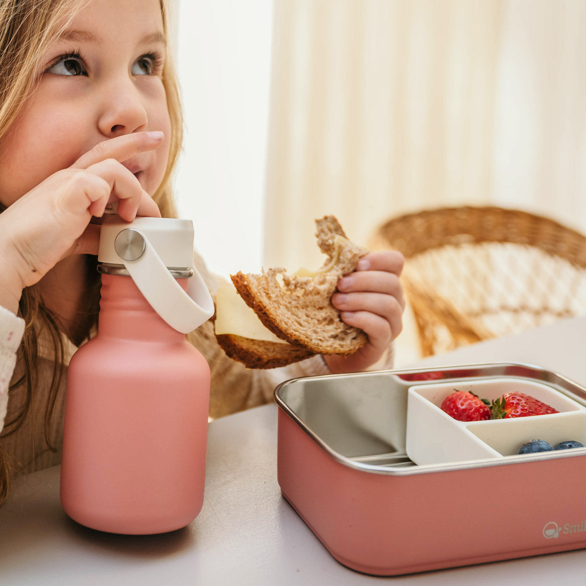 Meisje drinkt uit roze rvs fles en eet een boterham uit haar roze rvs lunchbox