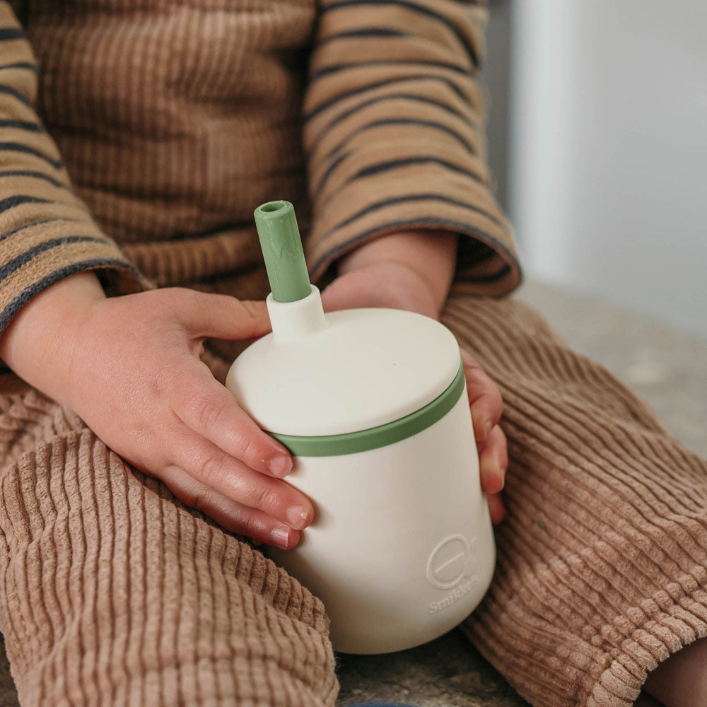 Handen van een kind houden een siliconen kinderbeker met groen rietje vast.