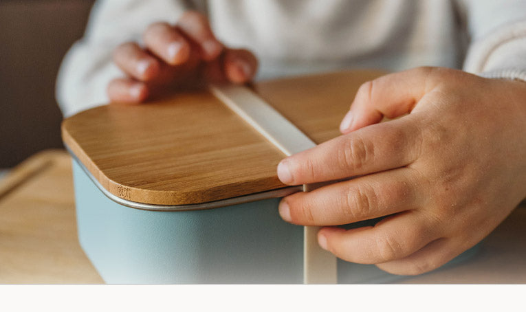 Jongetje houdt blauwe Smikkels lunchbox met bamboe deksel vast. De lunchbox heeft een witte elastiek