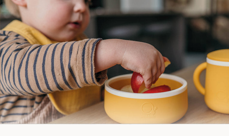Baby met gele slab haalt een stukje appel uit een geel siliconen kommetje van Smikkels