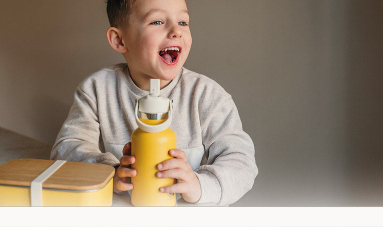Jongetje houdt een gele thermos drinkfles vast met daarnaast een lunchbox in dezelfde kleur met bamboe deksel