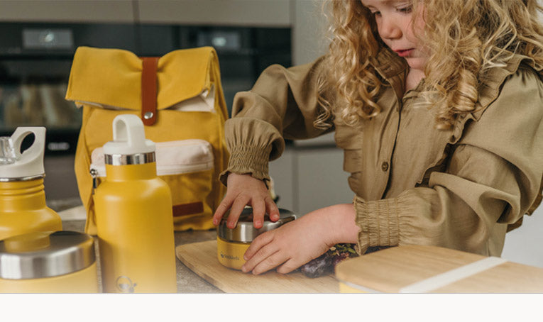 Meisje draai geel fruitbakje dicht met om haar heen drinkflessen, rugzak en lunchbox in dezelfde kleur van Smikkels