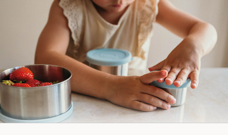 Meisje opend Smikkels RVS fruit- en snackbakjes met blauwe siliconen deksel. In de grootste zitten fris rode aardbeien