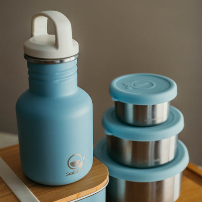 Smikkels drinkfles met witte dop, op een lunchbox met daarnaast drie RVS snack- en fruitbakjes met blauwe deksels