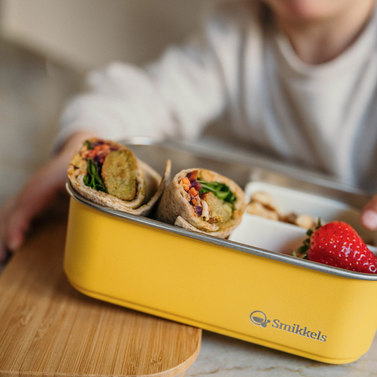 Kind houdt gele RVS lunchbox van Smikkels vast met wraps en aarbei