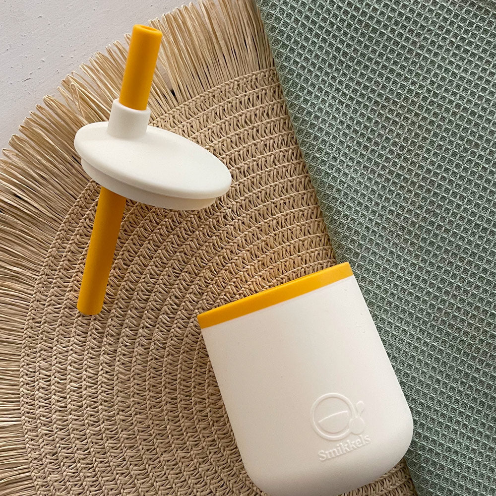 Sfeer en detail foto van een witte siliconen Smikkels drinkbeker. Ernaast de witte deksel met een geel rietje.