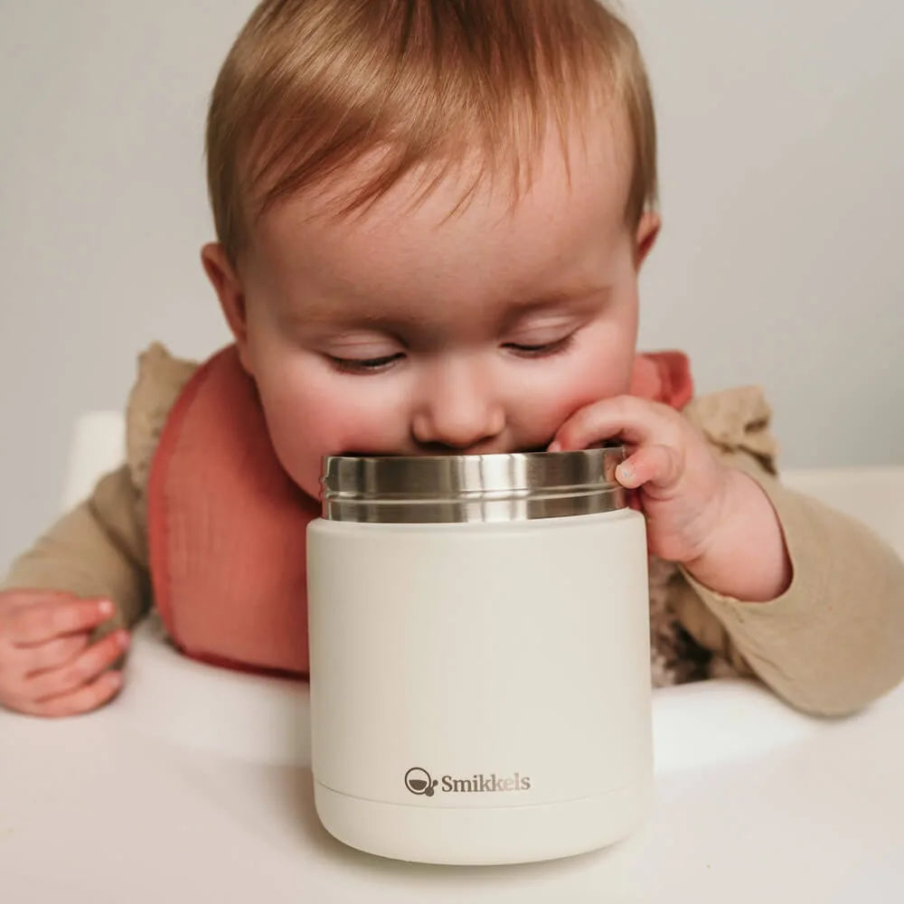 Baby met roze slabbetje in kinderstoel kijkt in wit RVS thermos lunchbakje van Smikkels